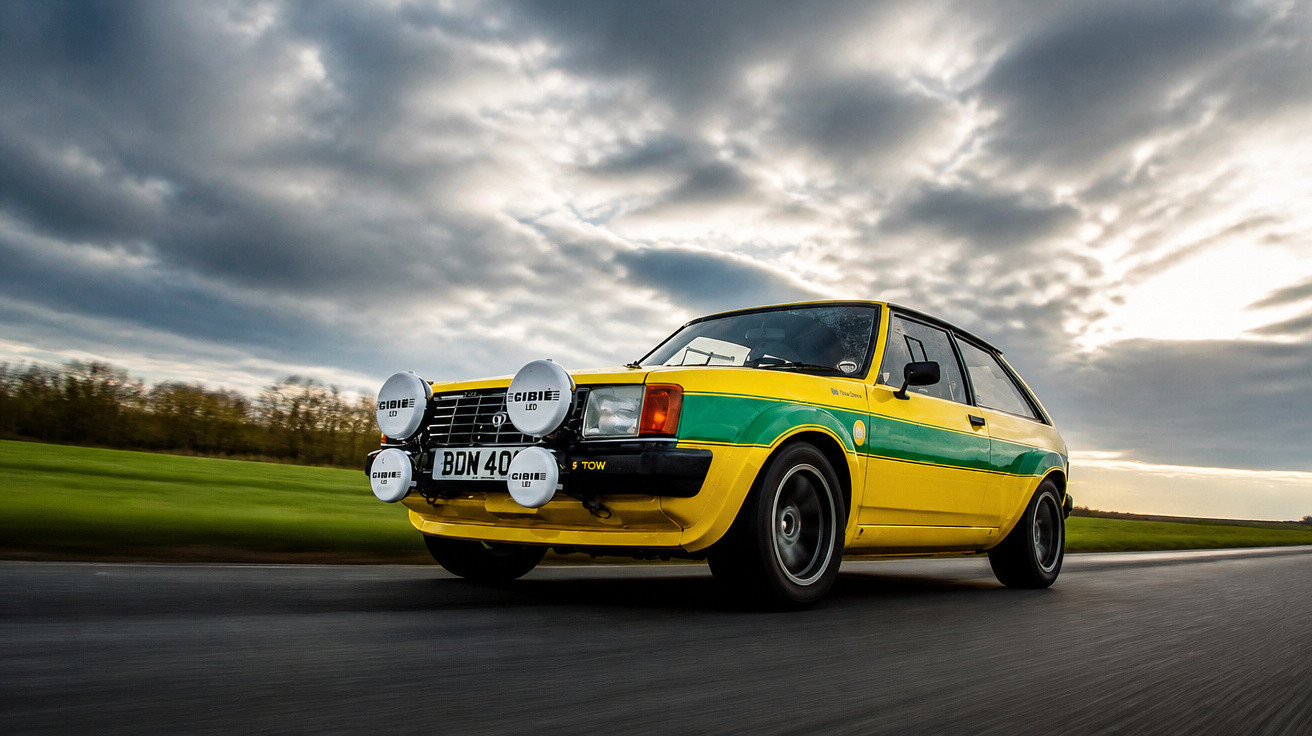 Voiture de rallye jaune à bande verte roulant sur route sous ciel nuageux en fin de journée.