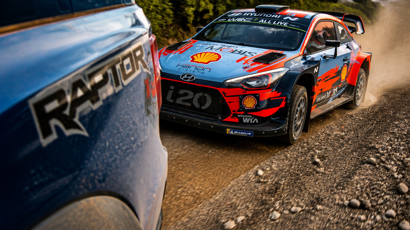 Voiture de rallye Hyundai i20 sur une route de terre en course, vue partielle d'un pick-up Ford Raptor.