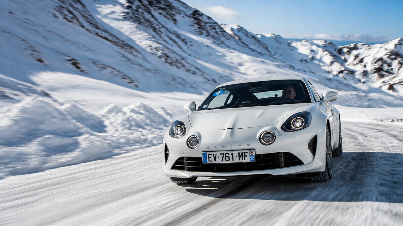 Voiture de sport blanche Alpine roulant sur une route enneigée en montagne sous un ciel bleu.