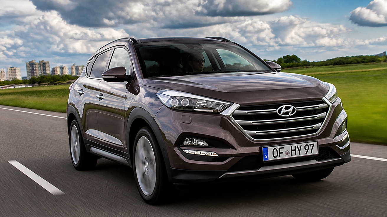 SUV Hyundai gris roulant sur une route avec paysage vert et ciel nuageux en arrière-plan.