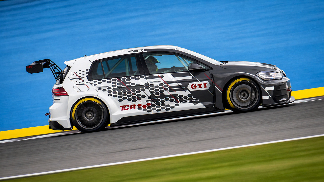 Voiture de course GTI blanche et noire sur circuit avec arrière aileron, pneus slicks et décor hexagonal.